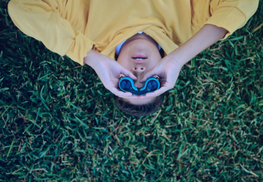 A kid laying in the grass holding binoculars.